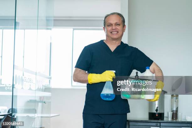 latino elderly office cleaner carries a basket of cleaning supplies in his hands while looking at the camera which lon portrays - office cleaning stock pictures, royalty-free photos & images