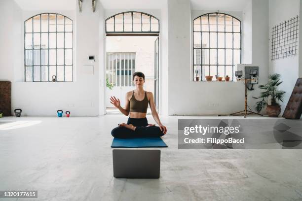 a young adult yoga teacher is waving her hand to her students on the laptop - yogi stock pictures, royalty-free photos & images