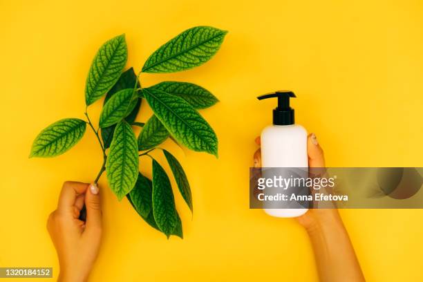 woman hands with manicure are holding green branch and white plastic bottle with dispenser on illuminating yellow background. trendy colors of the year 2021. flat lay style. copy space for your design - jojoba stockfoto's en -beelden