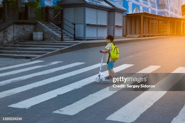 a boy with a backpack crosses the road on a zebra crossing on a scooter - cross road children stock pictures, royalty-free photos & images