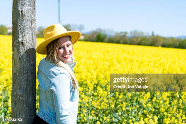 mujer rubia muy atractiva apoyada en un poste de madera en medio de un campo de flores amarillas. la mujer esta sonriendo y lleva puesto una chaqueta vaquera y un sombrero amarillo. estas flores se llaman colza o canola y se utilizan para hacer un tipo de - chaqueta de flores stockfoto's en -beelden