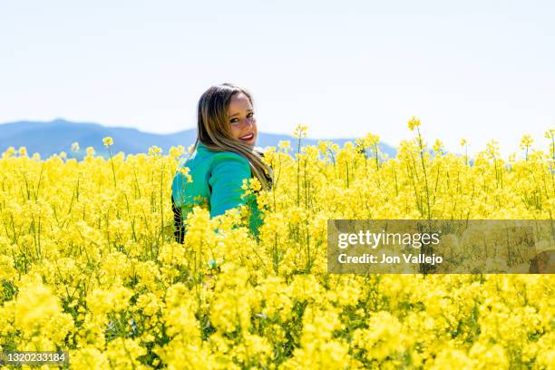 mujer rubia joven de perfil y mirando a camara en un campo de flores amarillas. lleva puesto una cazadora de cuero de color verde. estas flores se llaman colza o canola con la que se elabora un tipo de aceite. - canola oil stock pictures, royalty-free photos & images