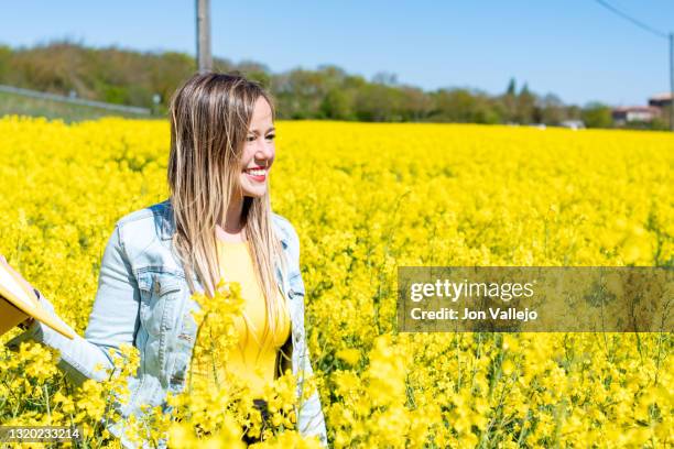 mujer rubia sonriendo entre muchas flores amarillas en un campo. estas flores se llaman colza o canola y se suelen utilizar para hacer un tipo de aceite. la mujer tiene un sombrero amarillo en la mano y lleva puesto una camiseta amarilla y una chaqueta va - chaqueta 個照片及圖片檔