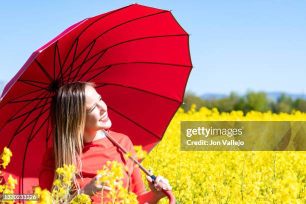 mujer rubia muy guapa esta sonriendo mientras mira al sol. lleva puesto un vestido rojo y lleva en la mano un paraguas rojo. ella esta en medio de un campo de flores amarillas llamadas colza o canola. - vestido rojo stock-fotos und bilder