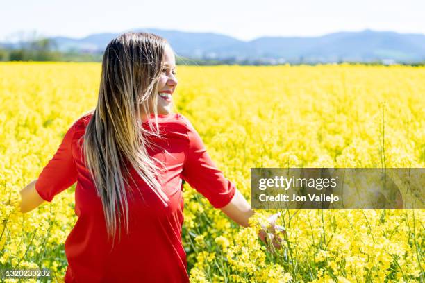 mujer rubia sonriendo con su vestido rojo mientras mira hacia su izquierda en medio de un campo lleno de flores amarillas. estas flores se llaman colza o canola y se suelen utilizar para hacer un tipo de aceite. - vestido rojo stock-fotos und bilder