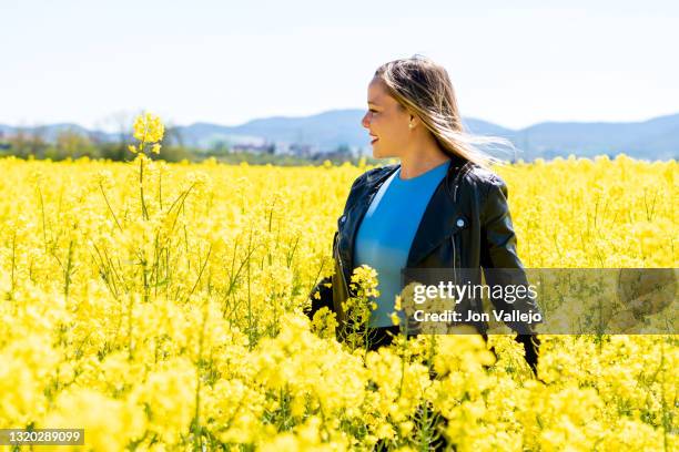 mujer rubia y joven entre flores amarillas. lleva puesto una cazadora de cuero negra y una camiseta azul. estas flores se llaman colza o canola con la que se elabora un tipo de aceite. - mujer negra stock pictures, royalty-free photos & images