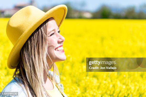 una joven mujer rubia muy guapa, esta sonriendo y mirando al sol en medio de un campo de flores amarillas. ella lleva puesto un sombrero amarillo y una chaqueta vaquera. estas flores se llaman colza o canola y se utilizan para hacer un tipo de aceite. - chaqueta stock-fotos und bilder