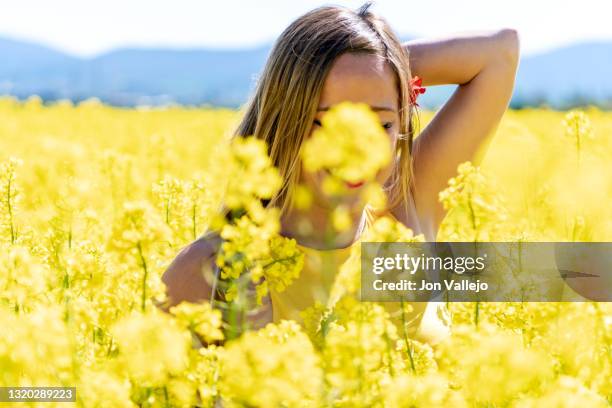 mujer rubia sonriendo con la mano detras de la cabeza esta entre muchas flores amarillas en un campo. esas flores se llaman colza o canola y se utilizan para hacer un tipo de aceite. ella lleva una flor roja en una oreja. - cabeza de flor stock-fotos und bilder