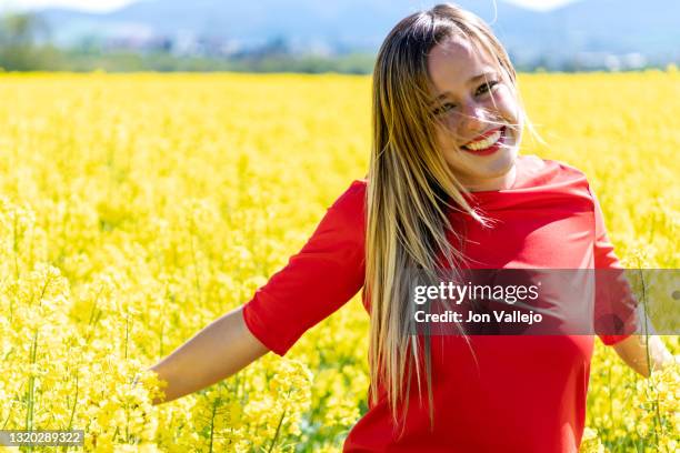 foto de medio cuerpo de una mujer rubia muy guapa que esta sonriendo con su vestido rojo en medio de muchas flores amarillas de un campo. estas flores se suelen utilizar para hacer un tipo de aceite y se llaman colza o canola. - vestido rojo stock pictures, royalty-free photos & images