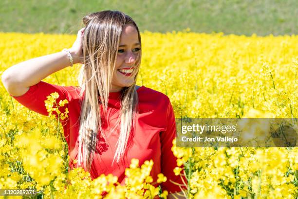 mujer rubia feliz sonriendo en medio de muchas flores amarillas. estas flores se llaman colza o canola. la mujer esta con la mano derecha sobre su cabeza, esta mirando hacia su izquierda y lleva puesto un vestido rojo. - vestido rojo stock-fotos und bilder