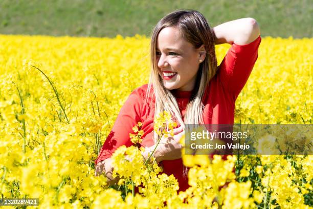 en medio de un campo de flores amarillas llamadas colza o canola, se encuentra esta bella modelo rubia sonriendo. la mujer lleva los labios pintados de rojo y lleva puesto un vestido de color rojo. - vestido de flores stockfoto's en -beelden
