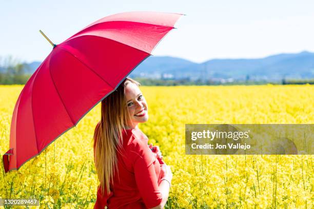 foto de medio cuerpo de una mujer rubia que esta sonriendo mientras pasea con su vestido rojo y su paraguas rojo por el medio de un campo de flores amarillas llamadas colza o canola. - vestido rojo stock-fotos und bilder