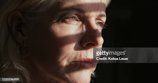 shot of a mature woman sitting alone in a dark room at home and looking outside contemplatively - close up face stock pictures, royalty-free photos & images