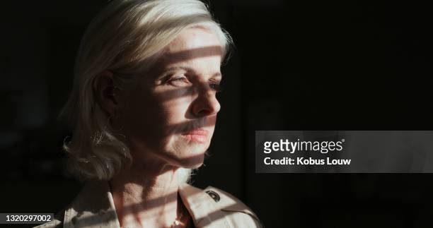 shot of a mature woman standing alone in a dark room at home and looking outside contemplatively - guilt stock pictures, royalty-free photos & images