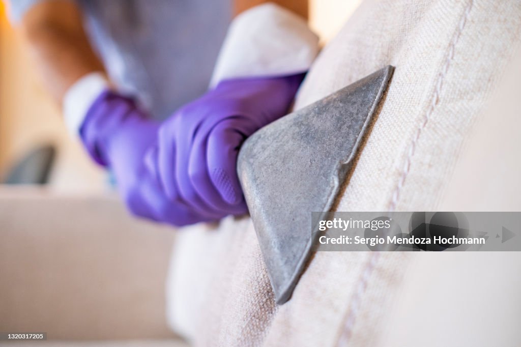Hand with a purple protective glove cleans a sofa's upholstery with a vacuum