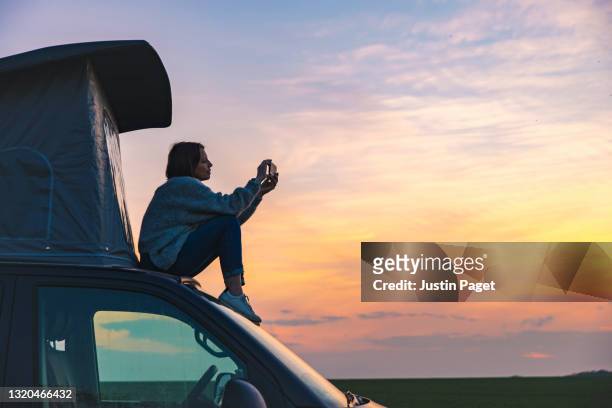 woman photographing the sunset from the rood of her campervan - reise stock-fotos und bilder