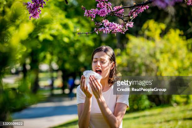 young pretty woman blowing nose in front of blooming tree. spring allergy concept - season stock pictures, royalty-free photos & images