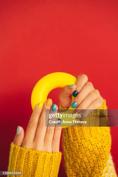 woman hands with bright manicure and in yellow knitted sweater are holding illuminating yellow bagel against red background. trendy colors of the year 2021. front view. copy space for your design - red nail polish stockfoto's en -beelden