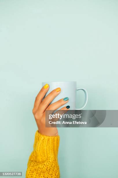 woman hand with bright manicure and in illuminating yellow knitted sweater is holding light blue cup against light blue background. trendy colors of the year 2021. front view. copy space for your design - pastellfarbig stock-fotos und bilder