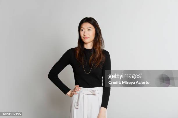 portrait of an asian millennial woman with long hair, smiling in front of a grey backdrop, wearing a black turtleneck and beige colored pants. - tre quarti foto e immagini stock