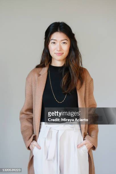 portrait of an asian millennial woman with long hair, smiling in front of a grey backdrop, wearing a camel colored coat, a black turtleneck and beige colored pants. - black pants woman fotografías e imágenes de stock