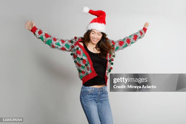 portrait of an asian millennial woman smiles while jumping in the air, wearing a  santa hat on her head, a red and green patterned ugly sweater and blue jeans. - ugly asian woman stock pictures, royalty-free photos & images