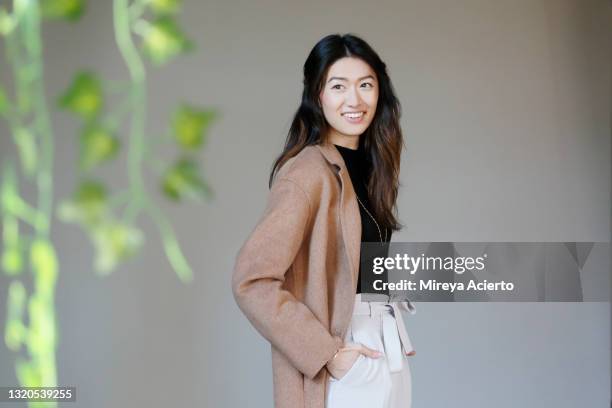 portrait of an asian millennial woman with long hair, smiling in front of a grey backdrop, wearing a camel colored coat, a black turtleneck and beige colored pants. - camel colored stock pictures, royalty-free photos & images