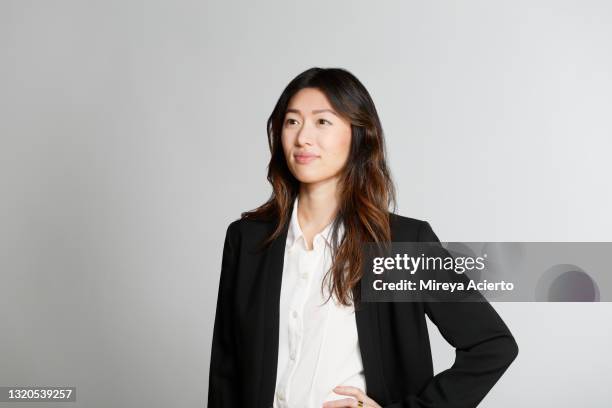 portrait of an asian millennial woman with long hair, smiling in front of a grey backdrop, wearing a black blazer, a white blouse and coral colored pants. - asian woman black shirt stock pictures, royalty-free photos & images