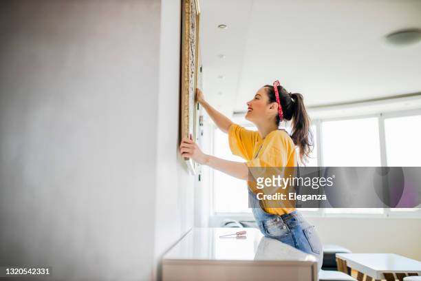jeune femme accrochant une photo à un mur avec un regard de concentration - accrocher photos et images de collection