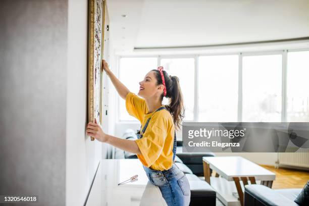 mujer joven colgando un cuadro en una pared con una mirada de concentración - decorating fotografías e imágenes de stock