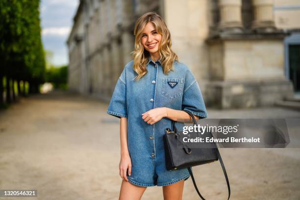 Xenia Adonts wears a Prada blue denim short jumpsuit with buttons and short flared sleeves, a black Prada bag, on May 27, 2021 in Paris, France.