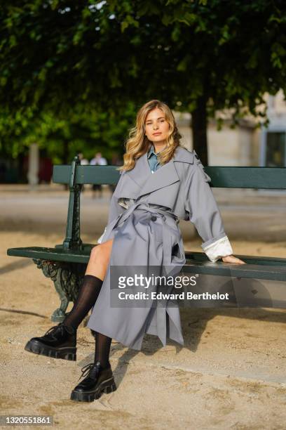 Xenia Adonts wears a Prada blue denim short jumpsuit with buttons, black long socks, black leather Prada shoes, a gray long trench coat from Attire...