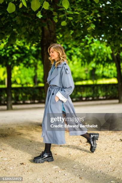 Xenia Adonts wears a Prada blue denim short jumpsuit with buttons, black long socks, black leather Prada shoes, a gray long trench coat from Attire...