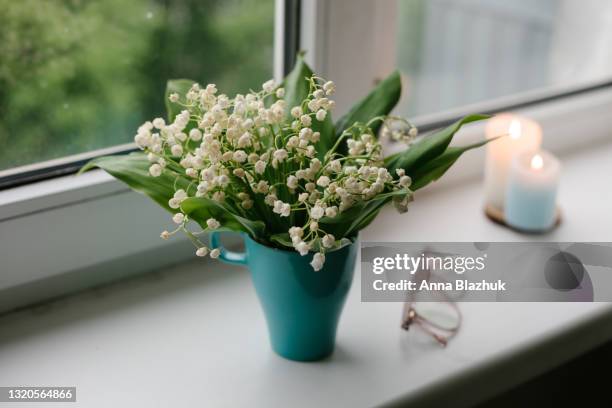 home decor or cozy home interior details. window sill with bunch of lily of valley spring white flowers in blue cup, eyeglasses and candles. - lily of the valley stock-fotos und bilder