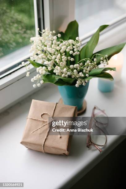 home decor or cozy home interior details. window sill with bunch of lily of valley spring white flowers in blue cup, eyeglasses and gift box wrapped with craft paper. - lily of the valley stockfoto's en -beelden