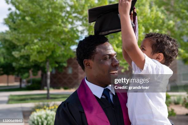 african american male college graduate in cap and gown holding and playing with his young three year old son on graduation day - son graduation stock pictures, royalty-free photos & images