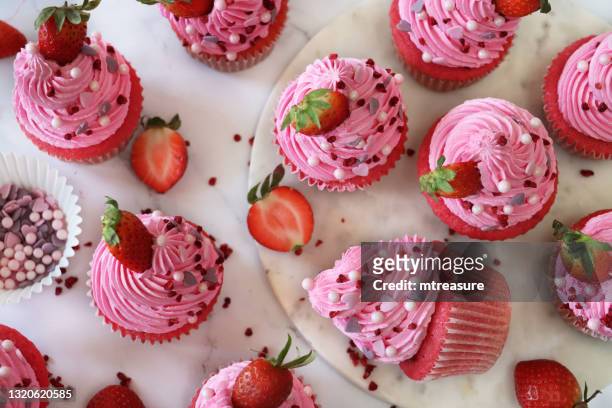 image of batch of homemade, pink velvet strawberry cupcakes in paper cake cases, strawberry on pink butter icing piped swirls, sugar craft heart-shape and sphere sprinkles, one cupcake lying on side, elevated view - cupcake stock pictures, royalty-free photos & images