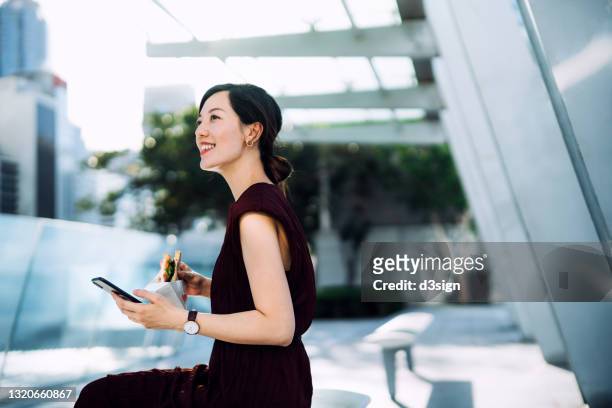 beautiful smiling young asian businesswoman sitting on the bench in office building terrace, using smartphone outdoors while eating a healthy sandwich during lunch break. lifestyle and technology - inspiring women luncheon ストックフォトと画像