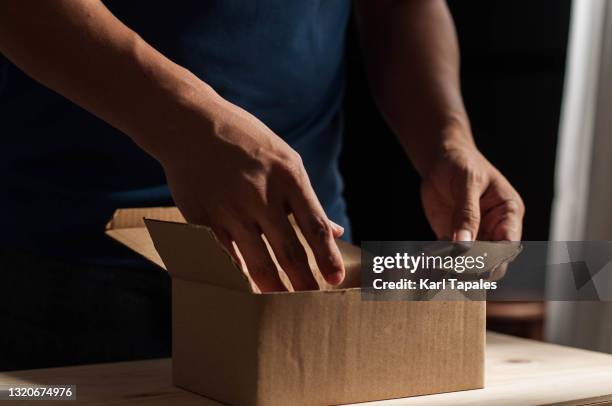 a young adult southeast asian man is unboxing a small carton box package at home - unboxing stock-fotos und bilder