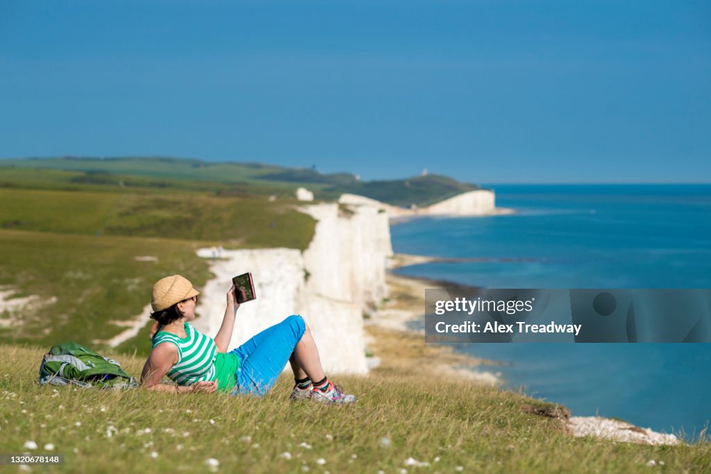 A woman reads her digital tablet on the cliffs nears Beachy Head with views of the Seven Sisters coastline in the distance