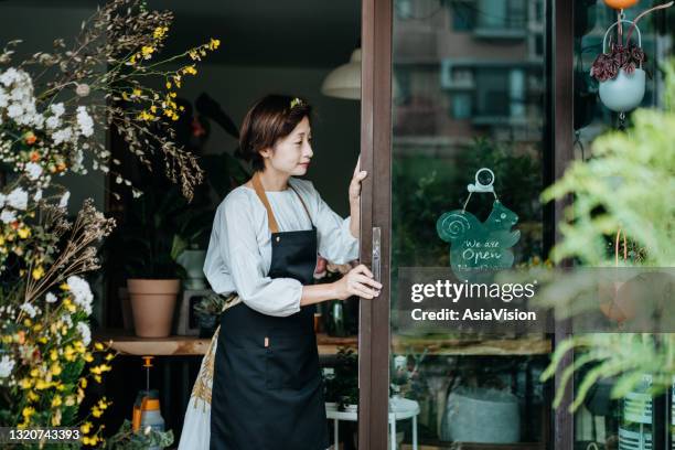 若いアジアの女性の花屋、彼女のビジネスの日を開始する中小企業の花屋の所有者。ドアを開けて、彼女の店で開いた看板をぶら下げる。中小企業のコンセプト。日常業務 - 開店 ストックフォトと画像