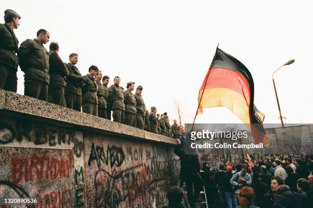 Jubilant man waves the German national flag under the gaze of East-German border guards on top of the Berlin Wall, Berlin, Germany, 10th November...
