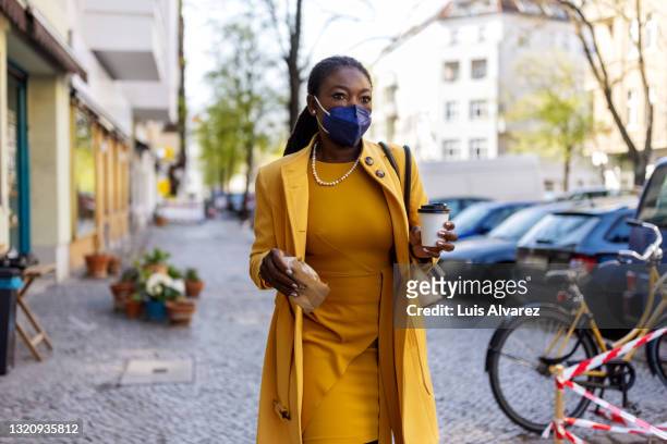 woman wearing mask walking in the city holding a take away food and coffee - abrigo amarillo fotografías e imágenes de stock