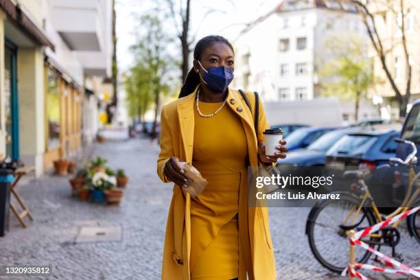 woman wearing mask walking in the city holding a take away food and coffee - yellow coat 個照片及圖片檔