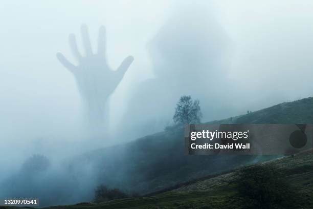 a spooky transparent hooded figure holding up it's hand. appearing over a moody hillside. on a foggy winters day in the countryside. - 巨人 ストックフォトと画像