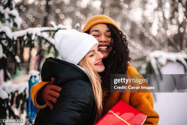 happy girlfriends embracing holding christmas gift by christmas tree - friends christmas foto e immagini stock