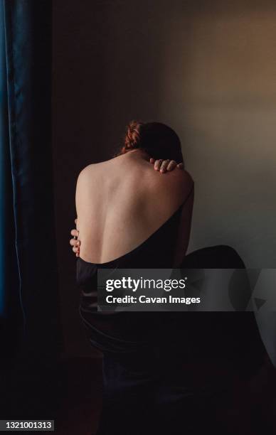 back view of sad woman bent forward with back exposed in a dark room. - woman head in hands sad stock pictures, royalty-free photos & images
