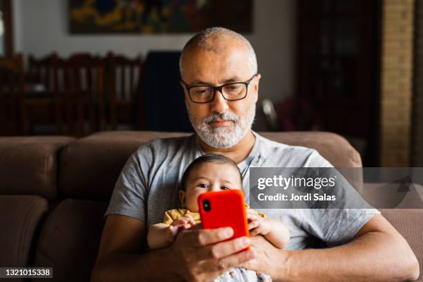 man sitting on a sofa watching an smartphone with his baby - baby mobile 個照片及圖片檔