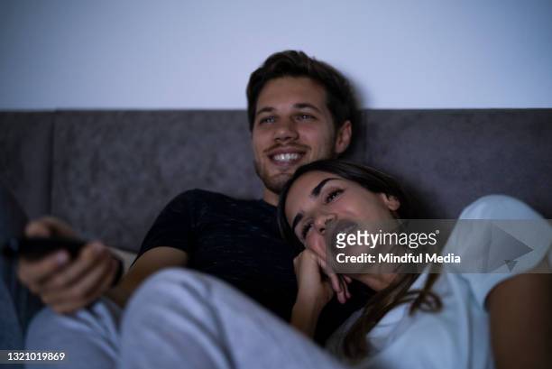 porträt eines fröhlichen paares, das auf dem bett liegt, während man im schlafzimmer fernsehen wird - zusehen stock-fotos und bilder