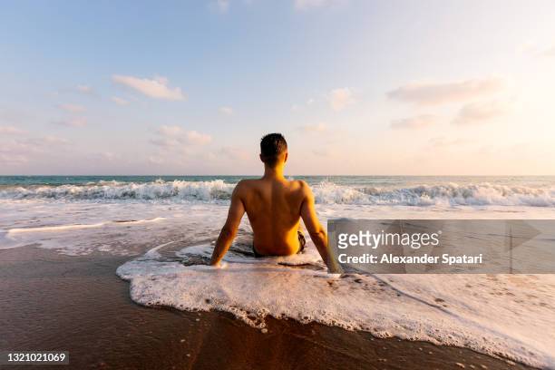 young man sitting at the beach and looking at the sea, rear view - beach sunbathing spain fotografías e imágenes de stock